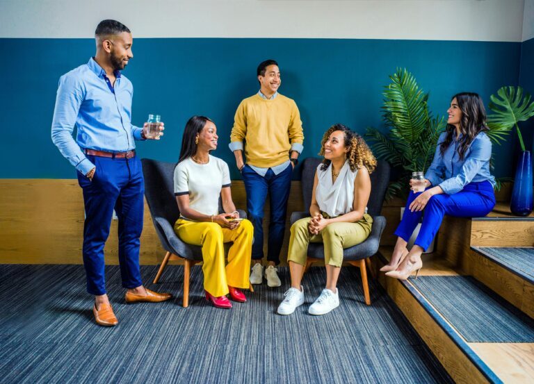 A diverse group of five professionals engaging in a casual indoor meeting, smiling and talking.